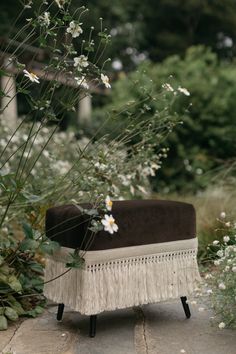 a chair with fringe trim and flowers on the ground in front of some bushes,