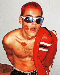 a shirtless man with red paint on his face and jacket over his shoulders, wearing sunglasses