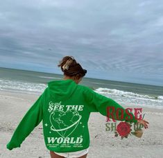 a woman is walking on the beach with her arms spread out towards the ocean while wearing a green shirt that says see the world