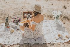 a woman sitting on top of a blanket next to a basket filled with fruit and flowers