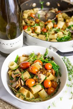 a white bowl filled with pasta and vegetables next to a bottle of wine