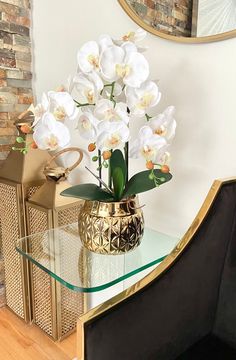 a vase with white flowers on a glass table next to a gold chair and brick wall