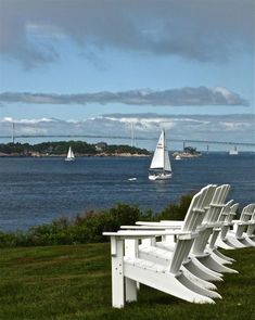 there are many white chairs on the grass by the water with sailboats in the background