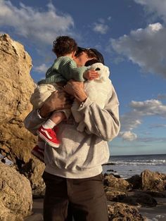 a man holding a small child on top of a rocky beach next to the ocean