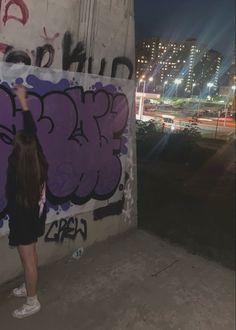 a woman is painting graffiti on a wall at night with buildings in the background and street lights