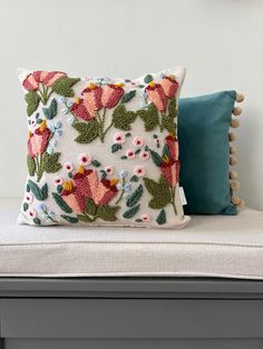 a white pillow with pink flowers and green leaves on it sitting on a gray bench