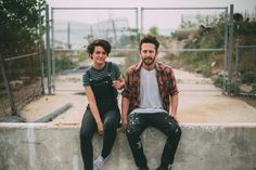 a man and woman sitting next to each other on a cement wall in front of a chain link fence