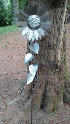 a metal sunflower sitting on top of a tree next to a dirt field with trees in the background