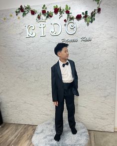 a young boy in a suit and bow tie standing on a white rug next to a sign