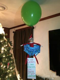 an elf is hanging from the ceiling in front of a christmas tree with a green balloon