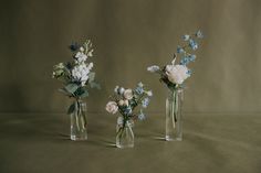 three glass vases with flowers in them on a brown tableclothed surface, one has blue and white flowers