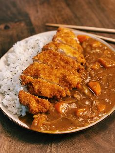 a white plate topped with meat and rice next to chopsticks on a wooden table