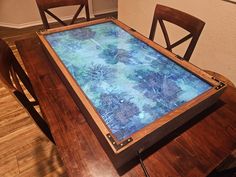 a wooden table topped with a glass top covered in blue and green flowers next to two brown chairs