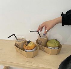 a person holding two cups with drinks and muffins in them on a table