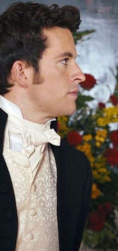 a man in a tuxedo and bow tie looking off into the distance with flowers behind him