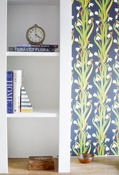 a book shelf with books and an alarm clock on it next to a wallpapered wall