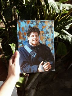 a hand holding up a photo of a man in blue and black with green leaves behind him