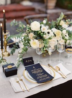 an elegant table setting with white flowers and black napkins, gold rimmed glasses and silverware