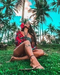 a woman sitting on the ground in front of palm trees with her eyes closed and hands behind her head