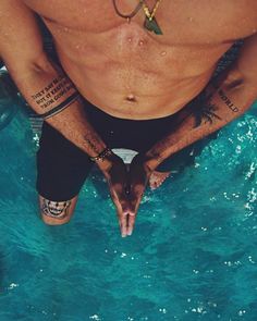 a man sitting in the water with tattoos on his arms and legs, looking down