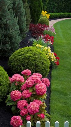 a garden with flowers and shrubs in the grass next to a white picket post fence