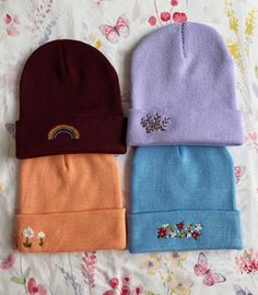 three beanies with embroidered rainbows on them are laying on a floral bed sheet