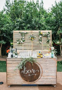 an outdoor bar with drinks and greenery on the top, sitting in front of some trees