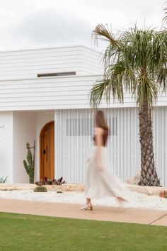 a woman in a white dress is walking past a palm tree and a large house