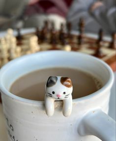 a cat figurine sitting on top of a coffee cup next to a chess board