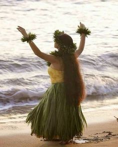 a woman in a grass skirt standing on the beach with her hands up to the sky