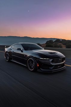 a black sports car driving down the road at dusk with mountains in the back ground