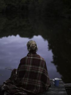 a woman sitting on the edge of a dock with her back to the camera, looking out at water