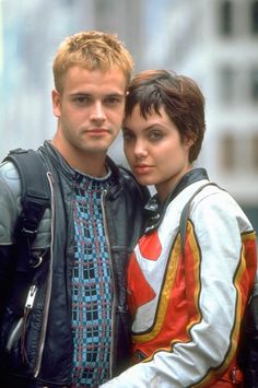 a man and woman standing next to each other in front of a tall building with buildings behind them