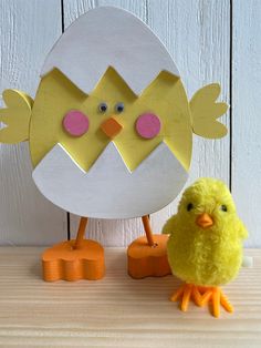 a yellow chick standing next to an egg on top of a wooden table with two smaller ones