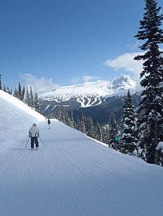 two people on skis going down a snow covered slope with mountains in the background