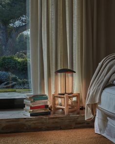 a bed sitting next to a window covered in curtains and books on top of a wooden table