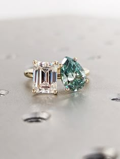 two engagement rings with green and white diamonds on top of a silver surface next to each other