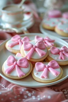 some cookies with pink bows on them are sitting on a white plate next to a cup and saucer