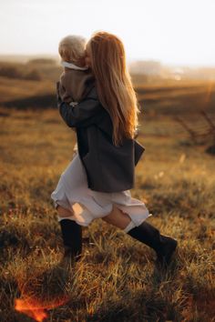 a woman holding a baby in her arms while walking through a field with the sun behind her