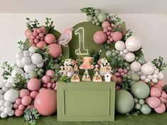 a green table topped with lots of balloons next to a wall covered in greenery