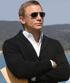 a man with his arms crossed standing in front of the ocean wearing sunglasses and a black cardigan