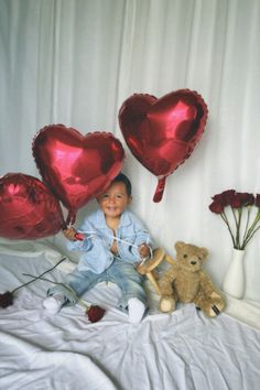 a young boy sitting on top of a bed next to balloons and a teddy bear