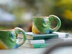 a green and yellow mug sitting on top of a book next to a cup that looks like an octopus