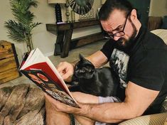 a man sitting on a couch reading a book with a black cat in his lap