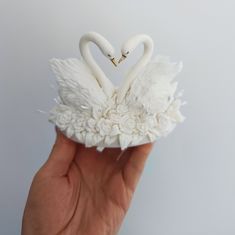 a hand holding a white swan ornament with two swans in it's beaks