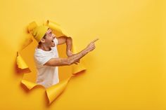 a man in white shirt and yellow hat holding his hand out with torn paper around him