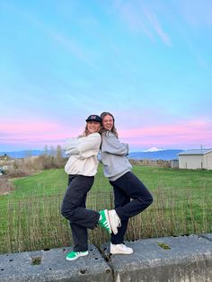 two women standing next to each other in front of a grassy field and blue sky