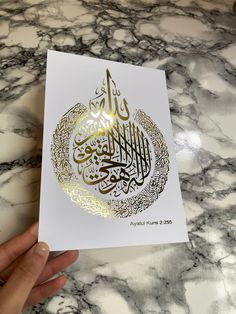 a hand holding up a card with an arabic calligraphy in gold on white paper