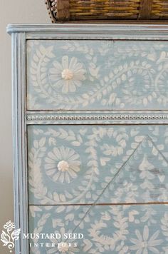 an old dresser painted in blue and white with flowers on the drawers, next to a wicker basket