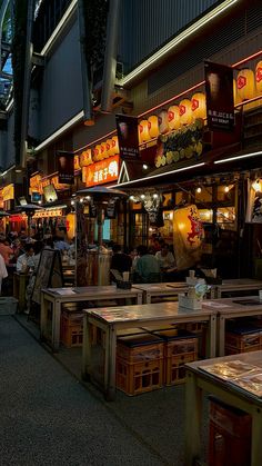 people are sitting at tables in an outdoor restaurant with neon lights on the building's roof